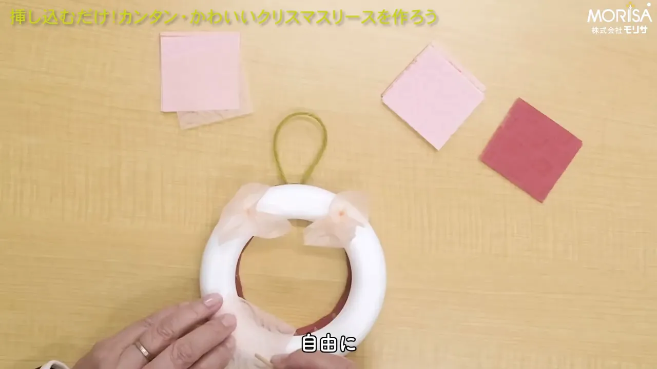 俯瞰で撮影された発泡スチロール製リースに和紙パーツがいくつか挿され、下側の手が配置を調整している様子。