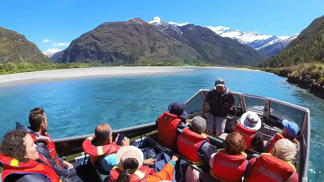 Tourists in a jetboat on a turquoise river with snow-capped New Zealand mountains and clear blue sky in the background.