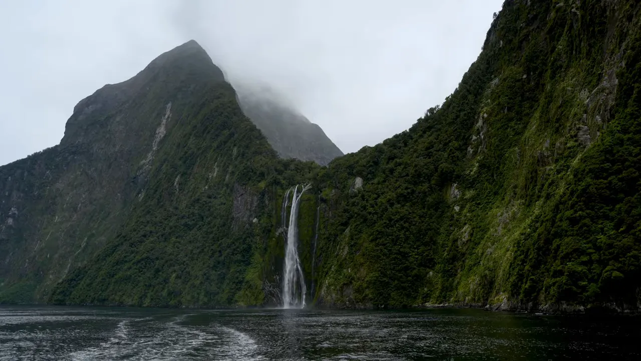 Wide view of Milford Sound showing a tall waterfall framed by steep, forested cliffs and the calm fjord water.