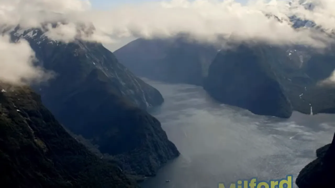 Aerial view of Milford Sound with low clouds, steep cliffs and a small boat on the water.
