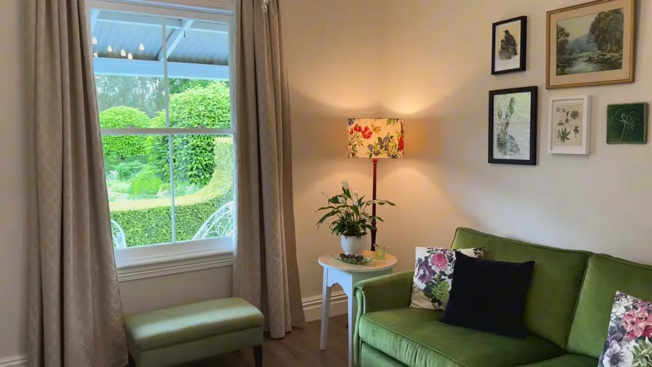 Cottage lounge with green sofa, floral cushions, ottoman and window looking to clipped garden
