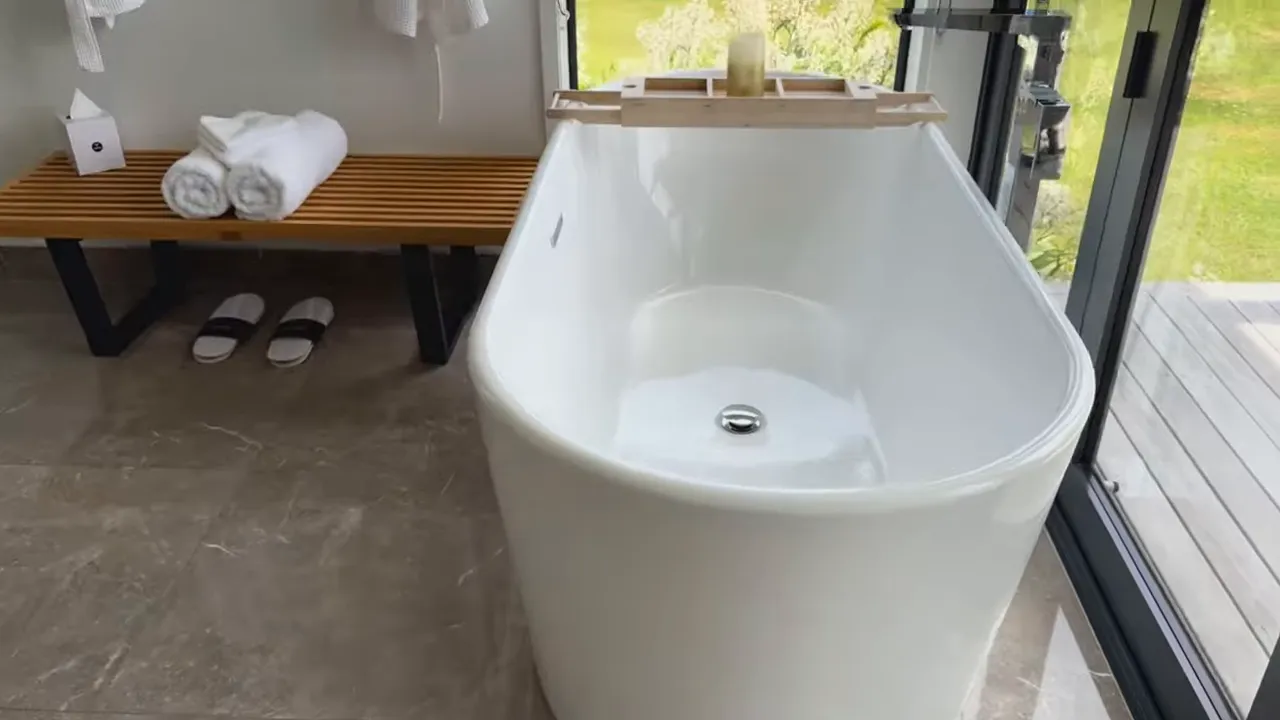 White freestanding bathtub with a wooden bath tray, towels on a bench and slippers on the floor beside a large window showing the garden.