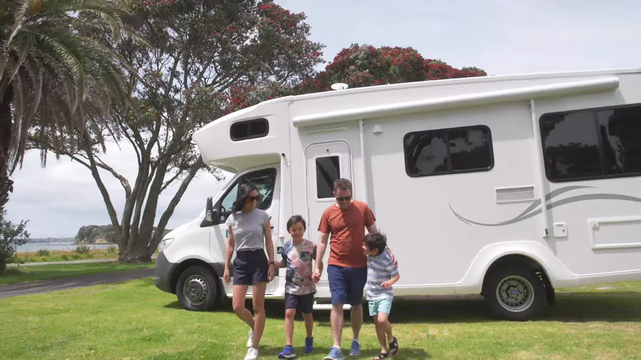 Family of four walking in front of a parked white motorhome on grass near trees and coastline