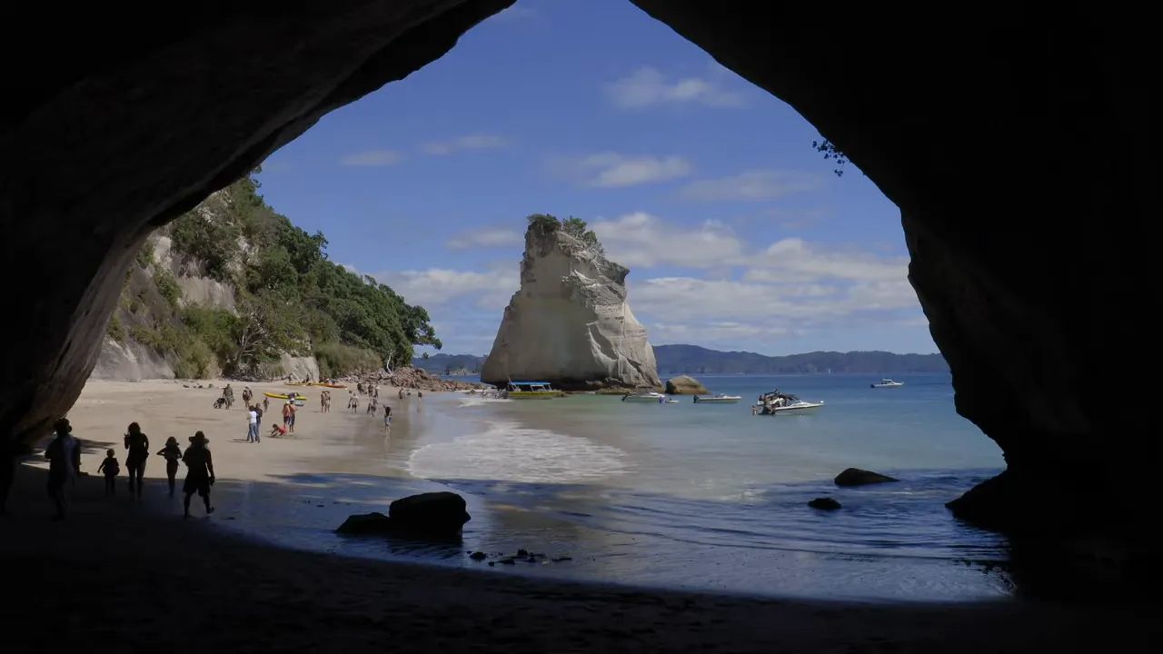 View from inside a sea cave looking out to a sandy beach, rock stack, boats and people at the shore