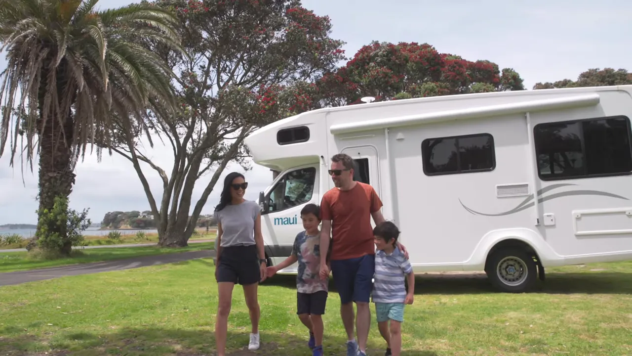 Family of four strolling in front of a white campervan parked by the coast with pohutukawa trees