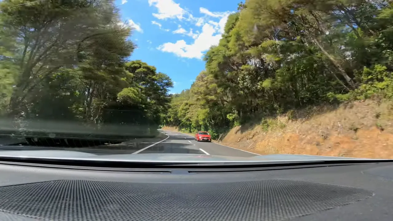 Driver's view from inside a car on a curving two-lane road with trees and a passing red car