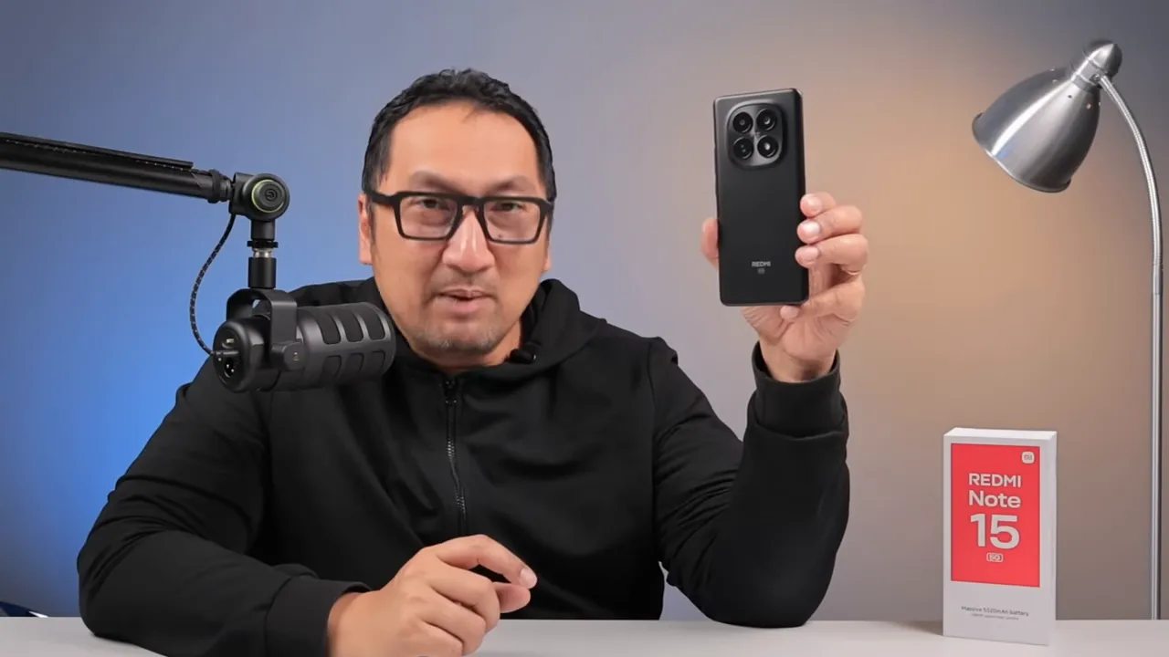 Reviewer memegang Redmi Note 15 5G di depan kotak penjualan pada meja, menampilkan desain dan modul kamera