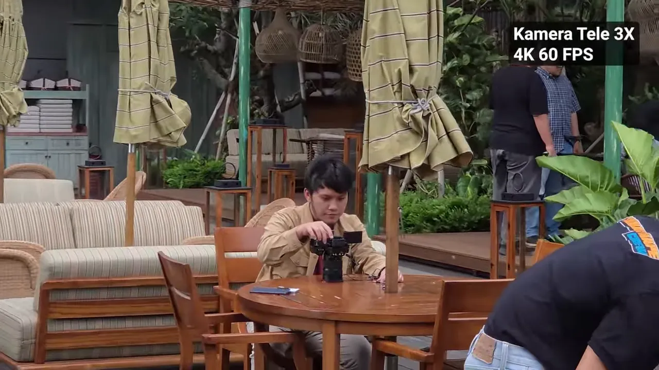 Close-up seorang pria di meja yang sedang menata kamera, dengan label 'Kamera Tele 3X 4K 60 FPS' terlihat di sudut.