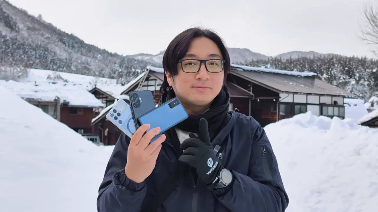 Tangan memegang tiga smartphone berjejer di depan latar rumah tradisional dan tumpukan salju