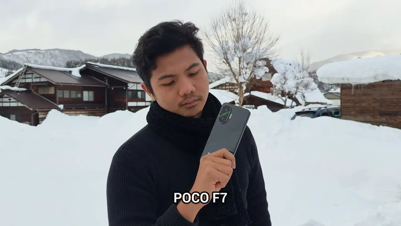 Penguji memegang POCO F7 di depan badan dengan teks identifikasi model di layar, latar lokasi bersalju