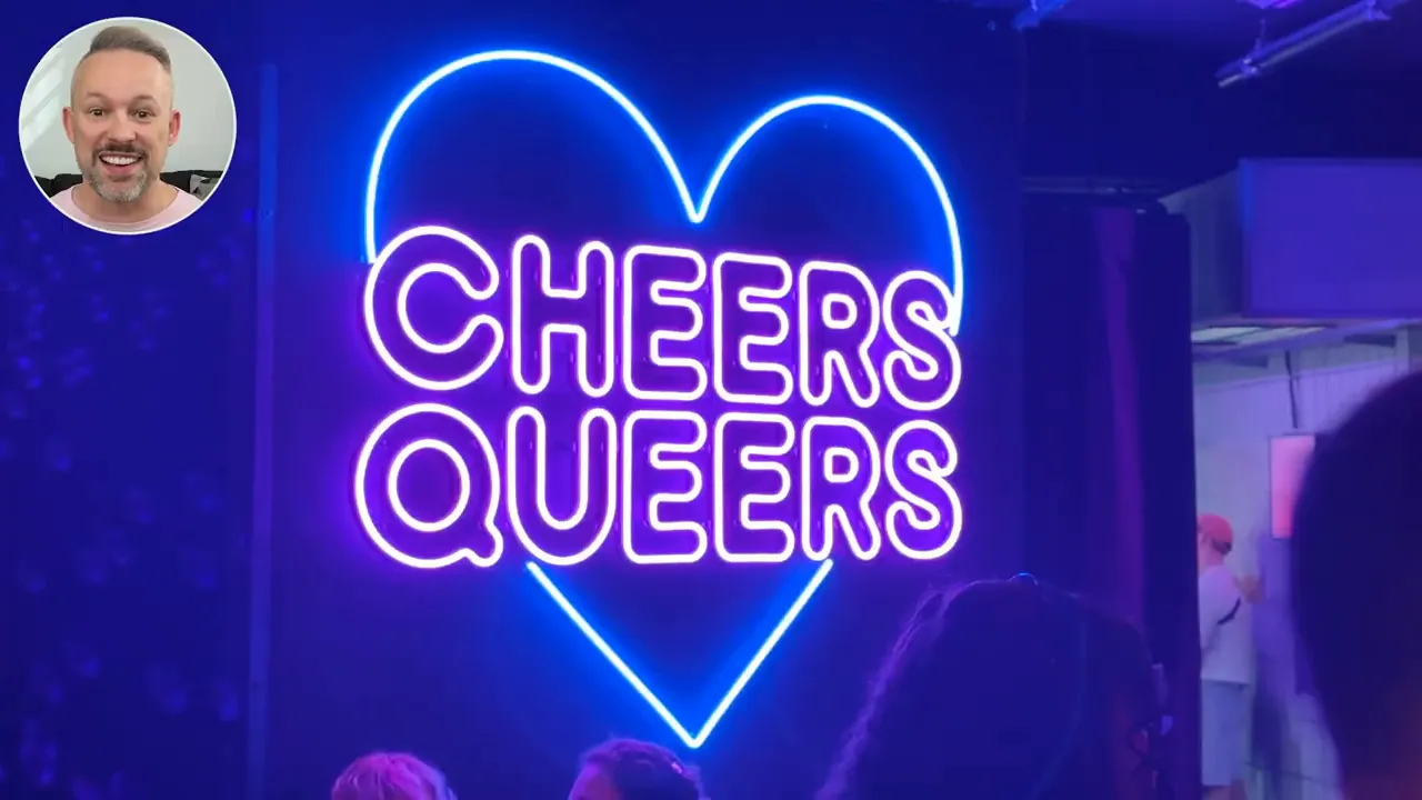 Bright blue and purple neon heart with the words 'CHEERS QUEERS' glowing on a club wall.