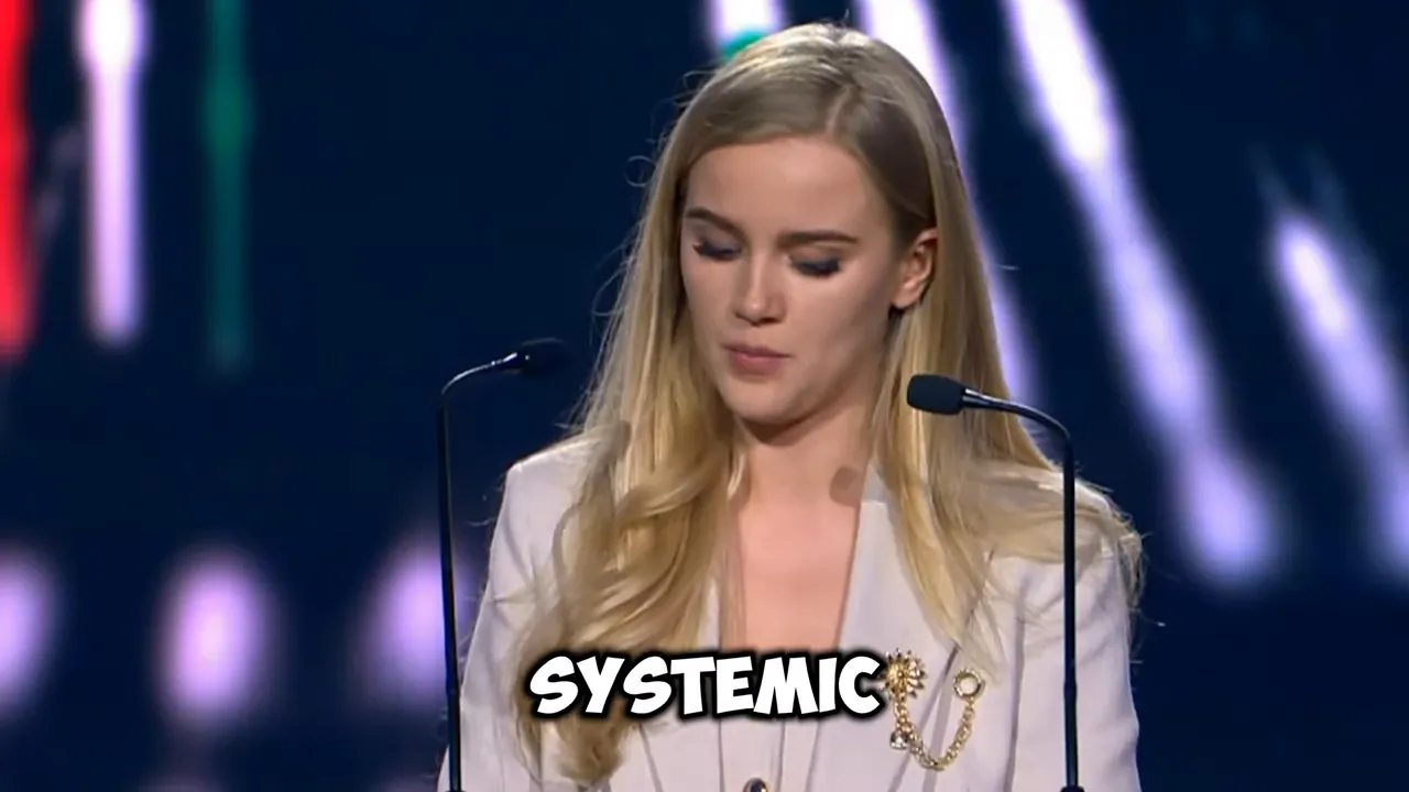 Eva Vlaardingerbroek speaking at a conference podium with microphones under stage lighting, with the word “SYSTEMIC” shown on screen