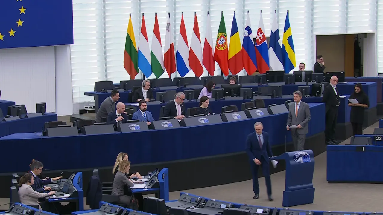 Wide view of the European Parliament plenary with rows of desks, national flags and officials.
