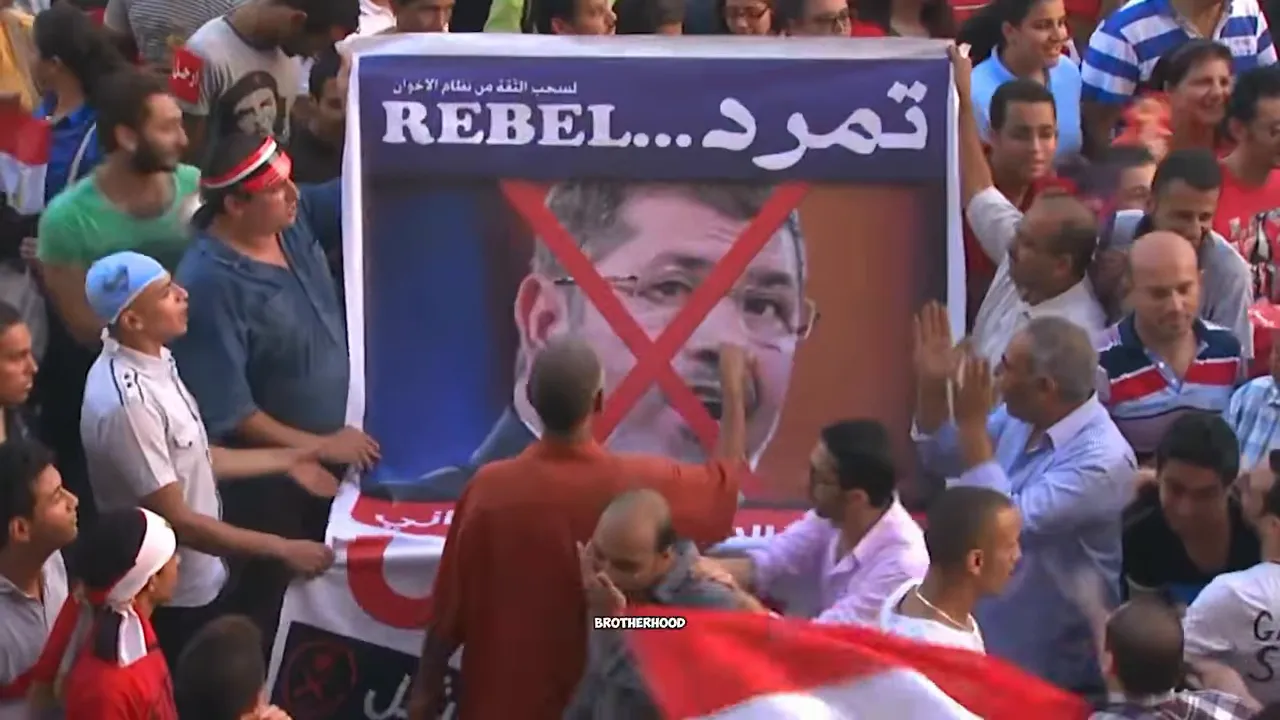 Large crowd holding a poster with a crossed out portrait and Arabic writing