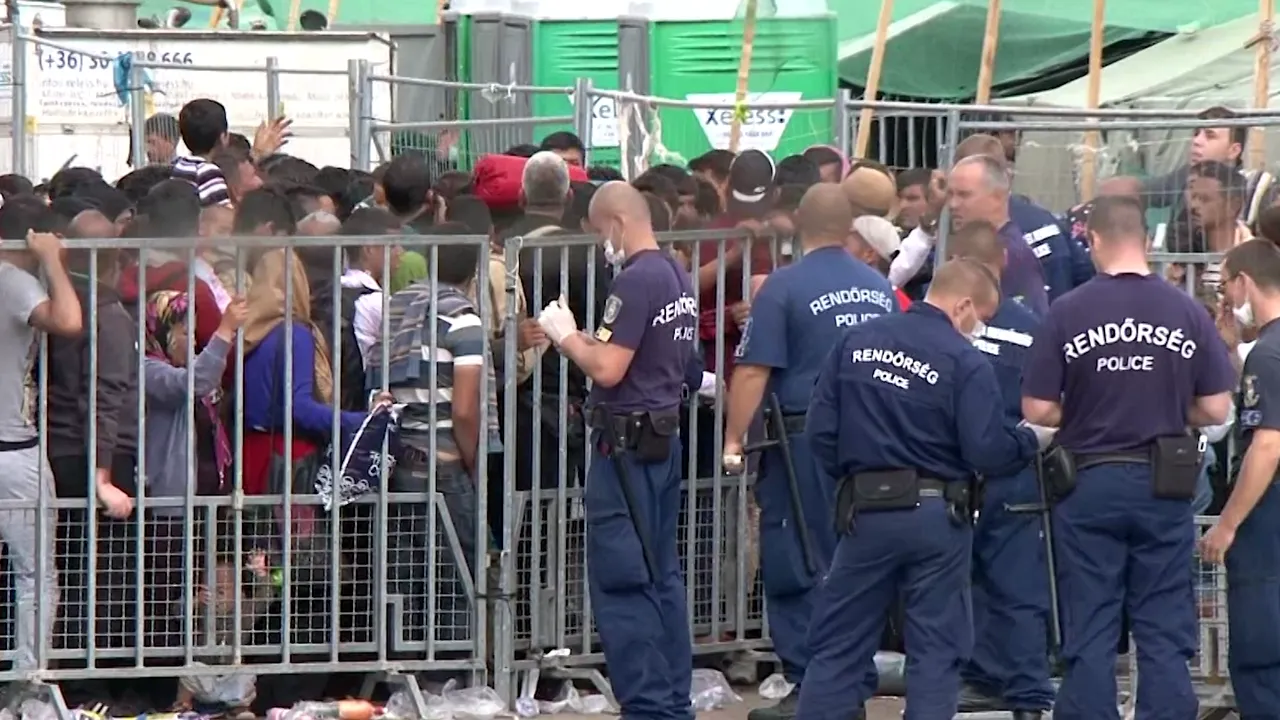 Crowded group of migrants pressed against metal barriers while police officers wearing 'Rendőrség' shirts manage the checkpoint.