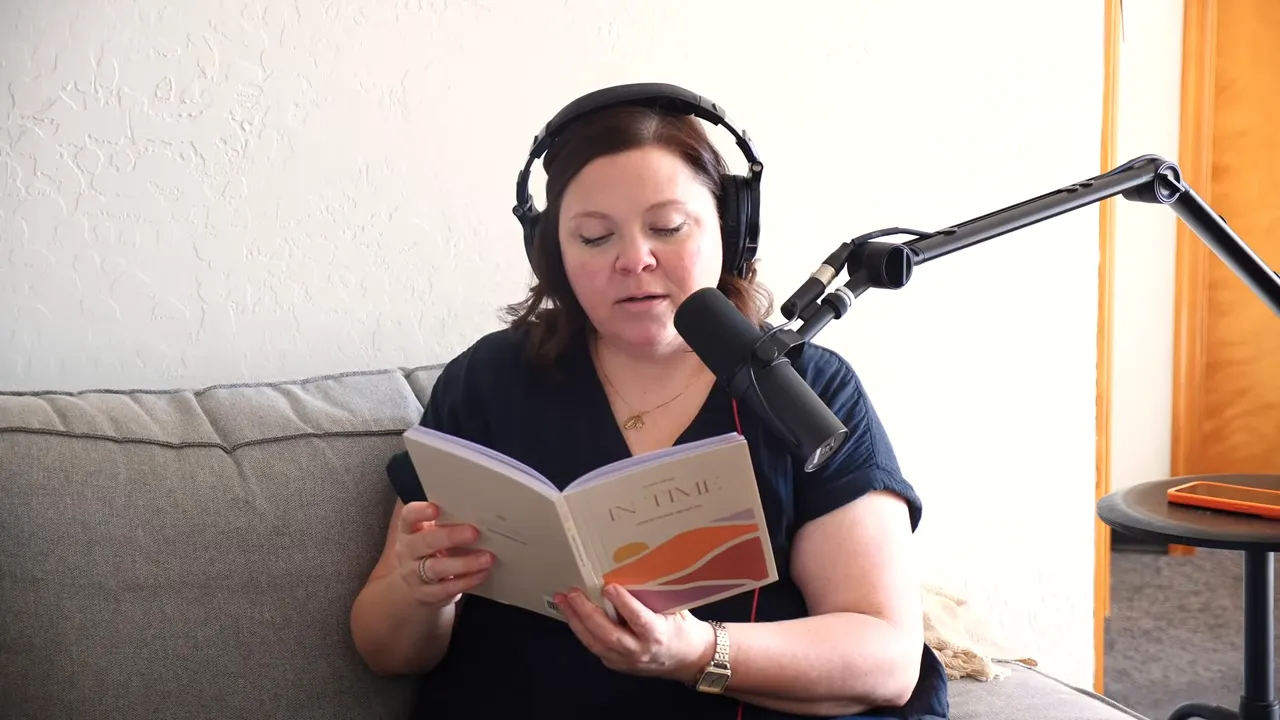 Person reading from a book into a microphone on a couch, wearing headphones, with an orange phone clearly visible on a nearby round side table.