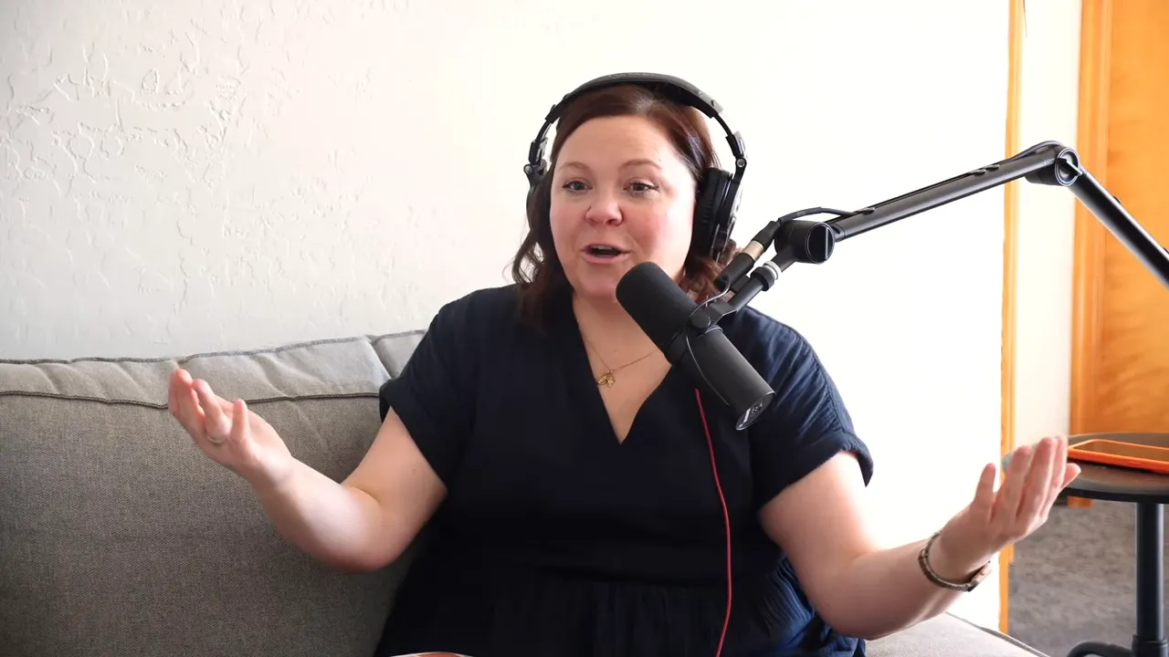 Podcast host seated on a couch wearing headphones and speaking into a microphone with arms open in a welcoming gesture.