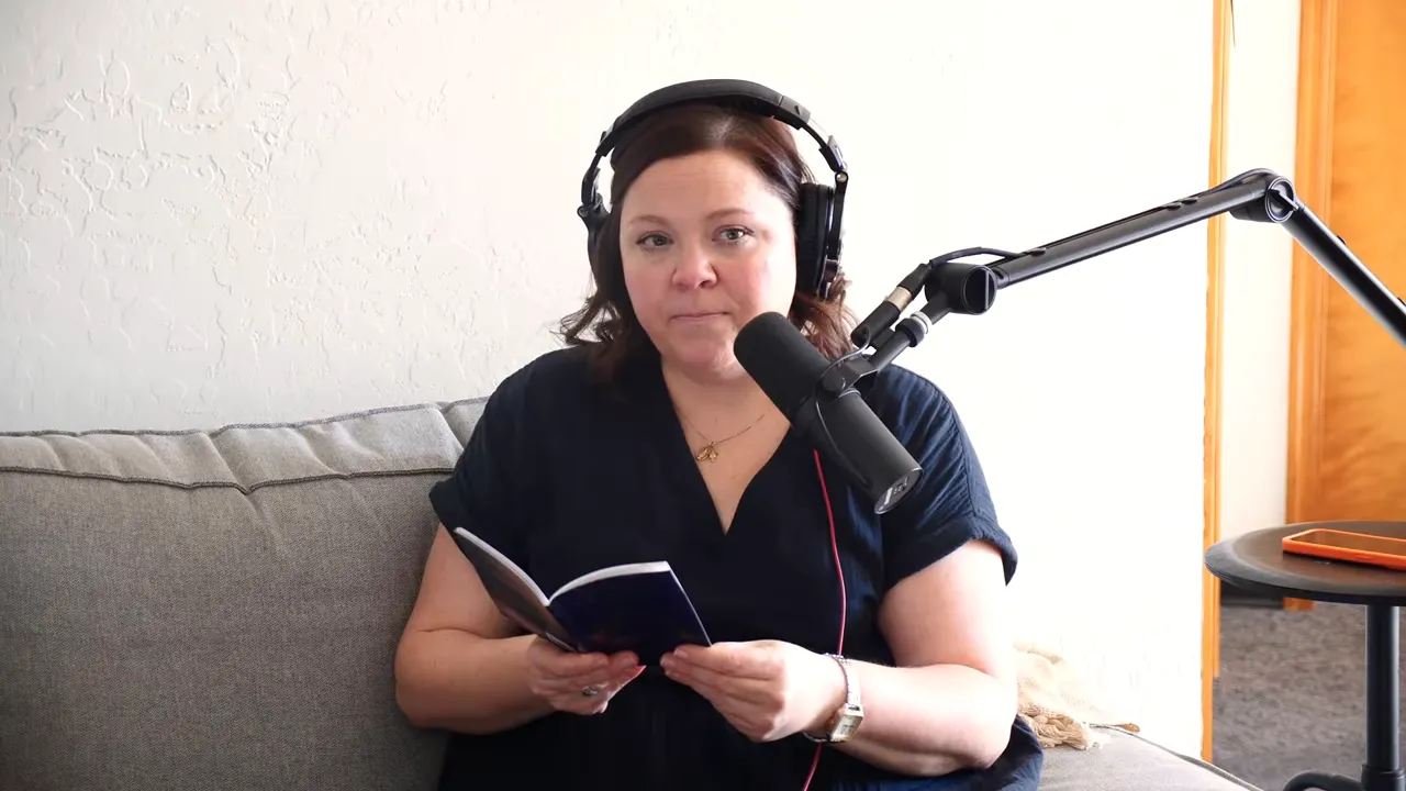 Podcast host looking directly toward the camera while holding a small book and speaking into a microphone, wearing headphones