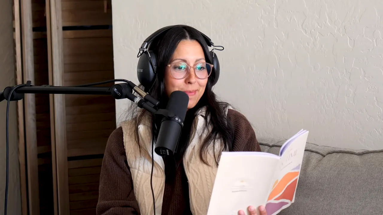 Podcast host wearing headphones and glasses, smiling softly while reading from a booklet with a microphone nearby, seated on a couch.