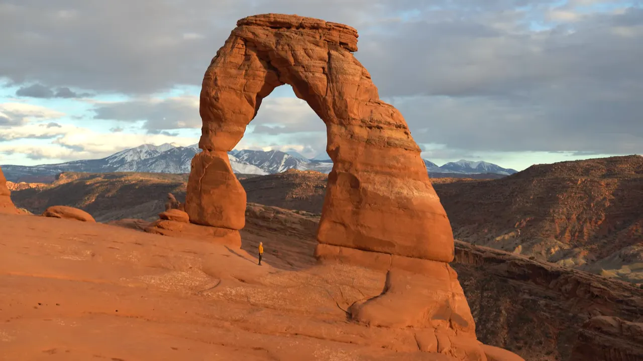 Delicate Arch glowing at sunset with La Sal Mountains in the background and a hiker for scale