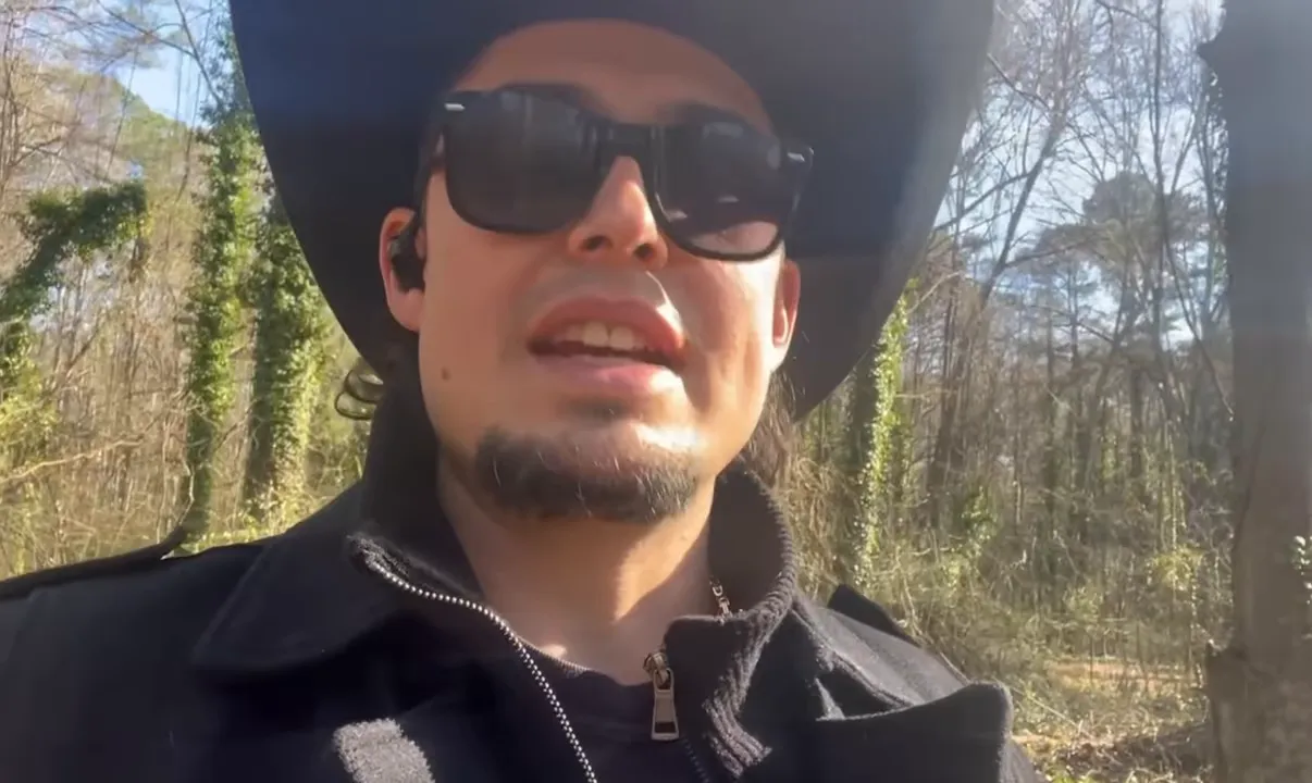 clear frontal shot of speaker in a cowboy hat and sunglasses speaking outdoors with trees behind