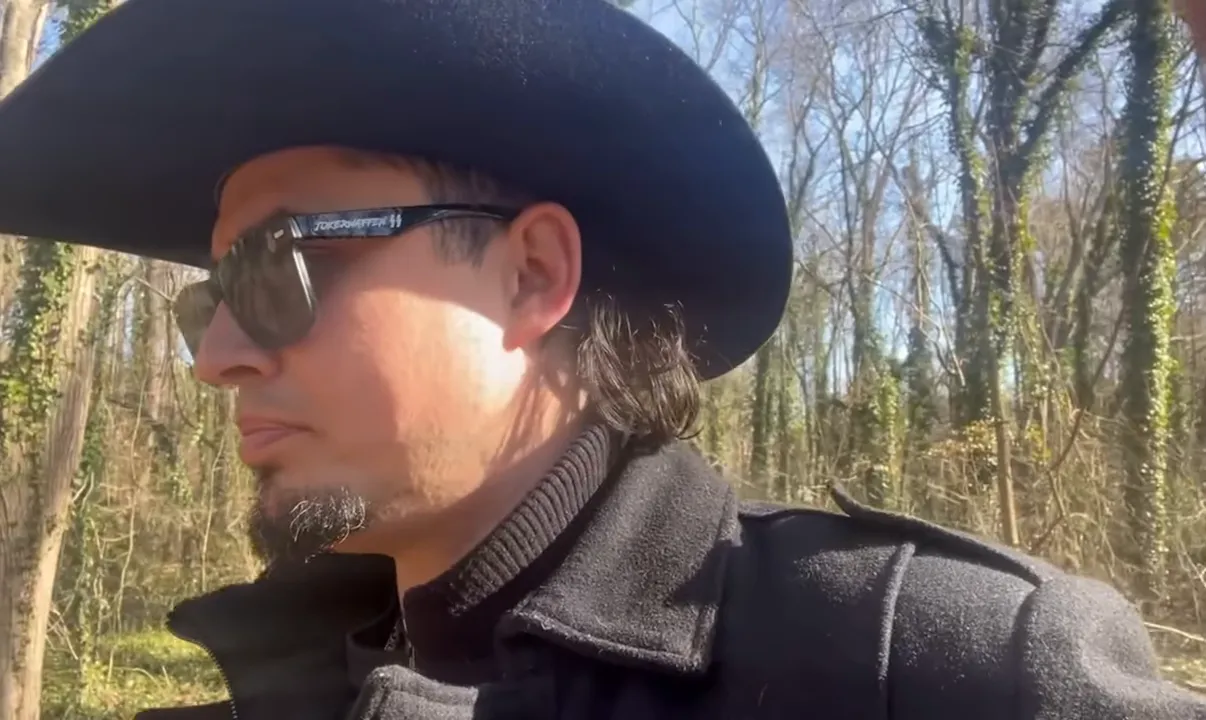 Speaker in a black cowboy hat gazing sideways in a sunlit forest, thoughtful expression