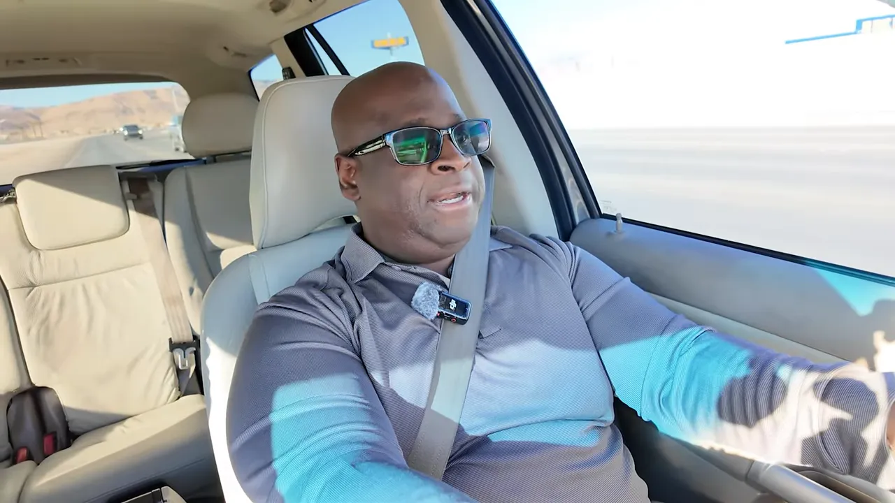 Presenter in a car, well-lit and centered, wearing sunglasses and a visible lapel microphone while speaking to camera.