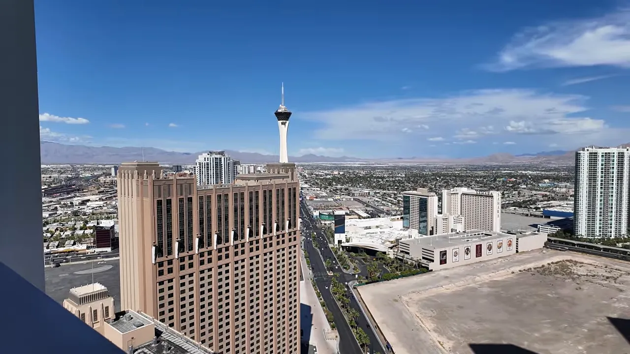 Wide north-Strip panorama from a Sky Las Vegas balcony showing the Stratosphere Tower, adjacent hotel towers and an empty parcel in the foreground.