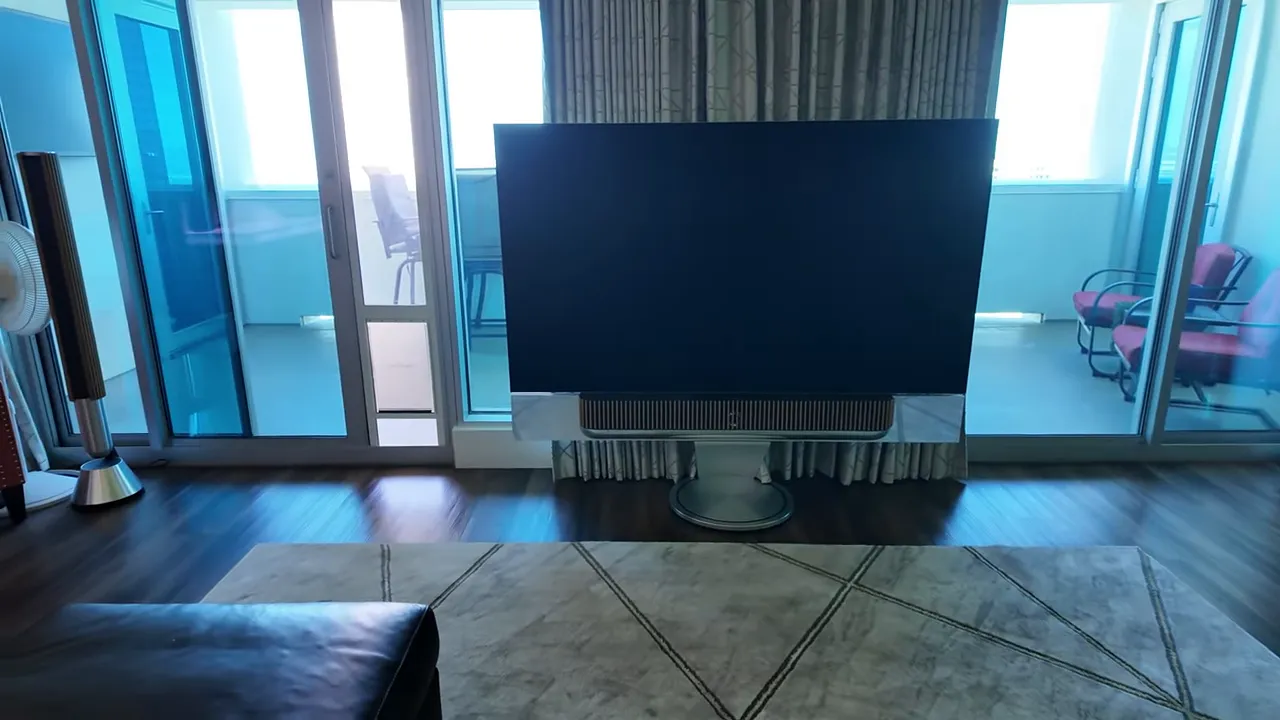 Front-facing shot of a large flat-screen television centered in the living room with floor-to-ceiling balcony doors behind it and a tower speaker to the side.