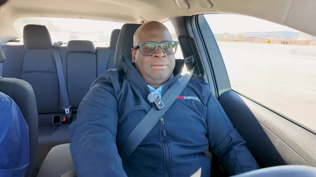 Wide, well-lit shot of the presenter seated in a car wearing sunglasses and a seatbelt, centered and speaking to camera.