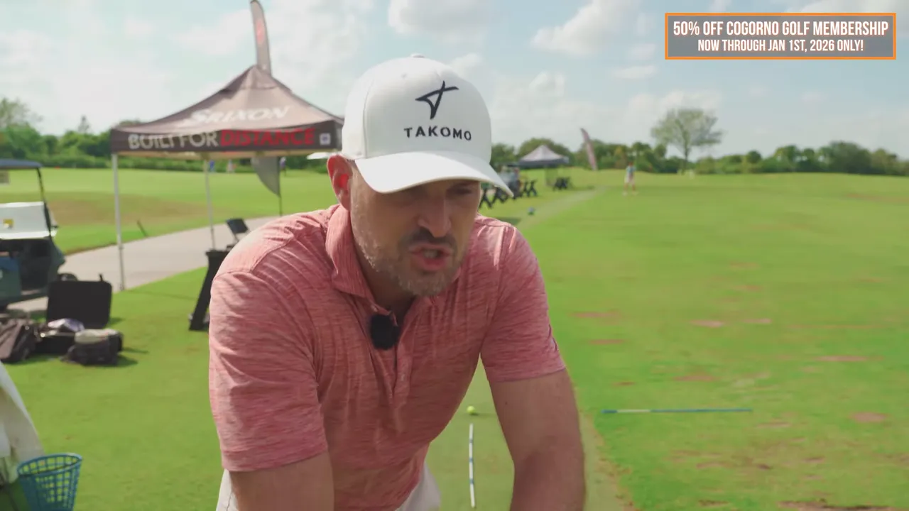 Instructor leaning forward toward the camera at the driving range delivering a coaching cue with alignment sticks on the mat behind him
