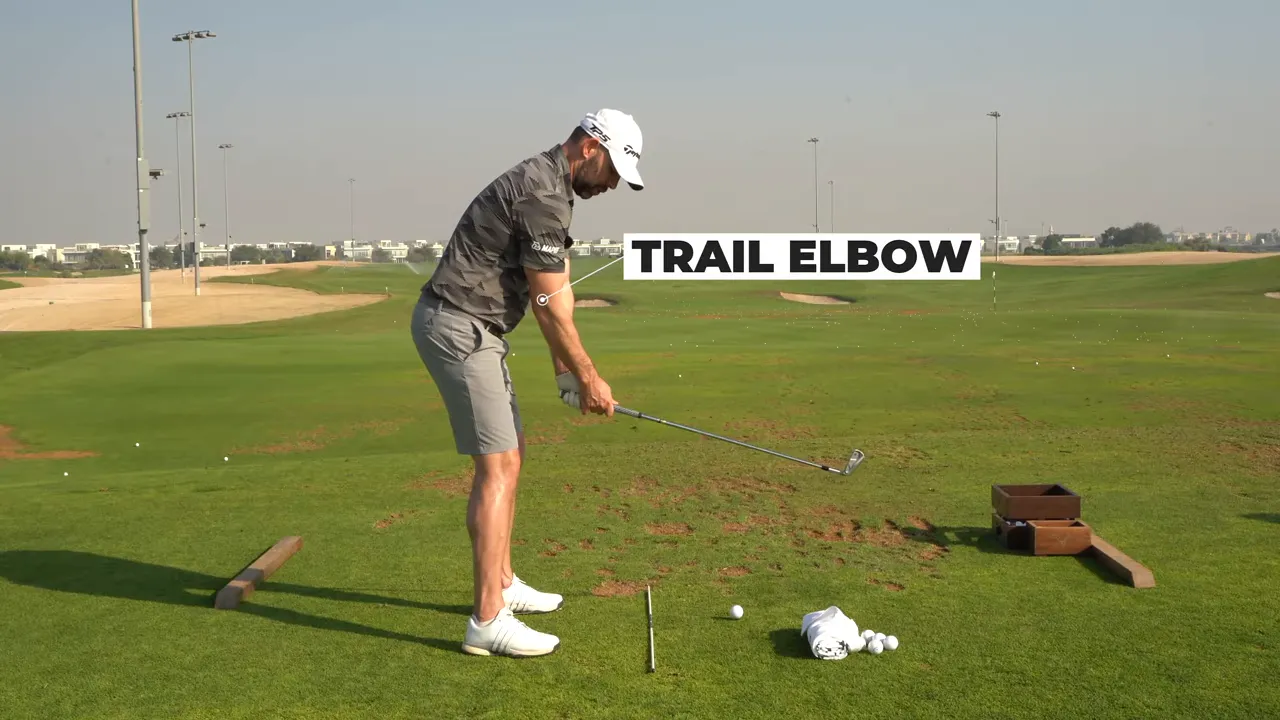 Wide shot of golfer at delivery with clear 'TRAIL ELBOW' label and alignment stick on turf.