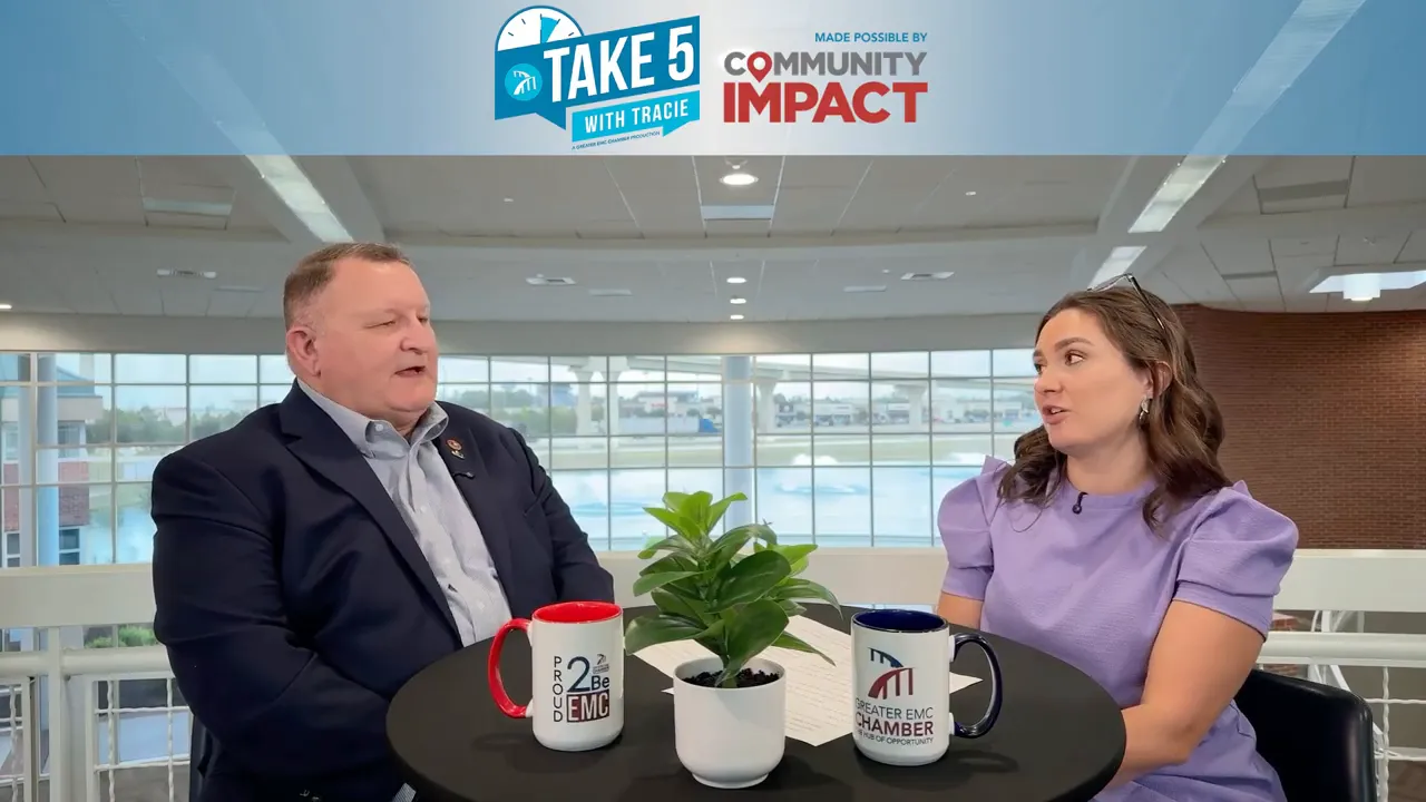 Wide interview shot showing host and guest at a round table with show branding, mugs and a potted plant