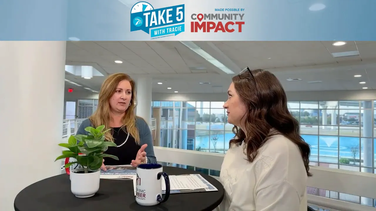 Profile view of two presenters in conversation at a round table with a plant, printed materials and a large windowed atrium behind them