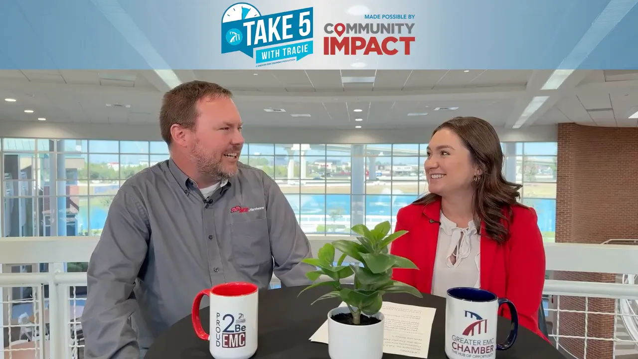 Host and guest in mid-conversation at a table with a plant and branded mugs during a recorded interview.