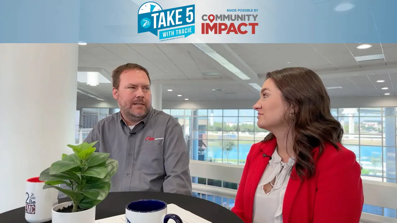 Two interview participants seated at a round table with mugs and a small plant in a bright atrium, mid-conversation.