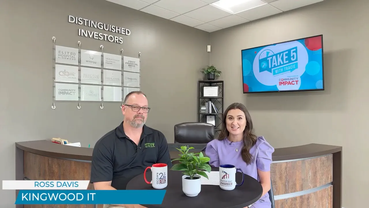 Two people seated at a small round table in an office lobby with mugs, a plant, and a wall mounted Take 5 screen behind them