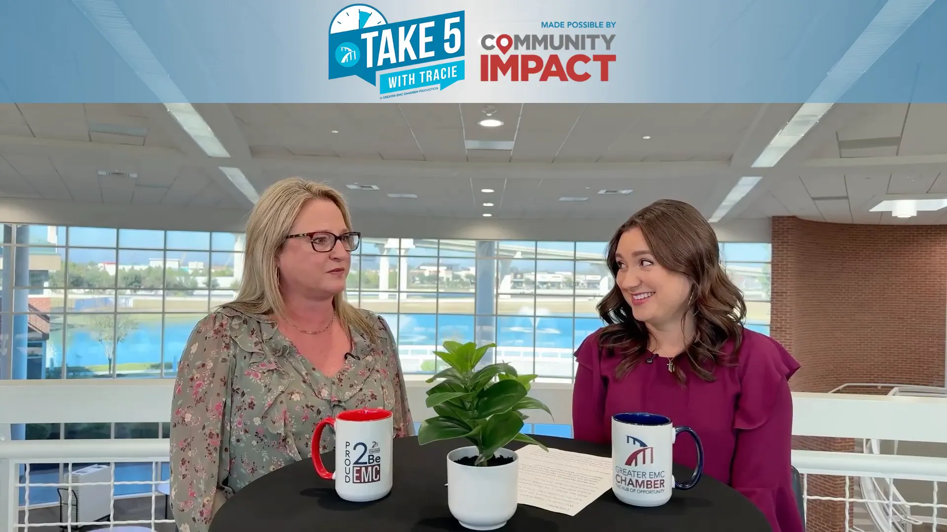 Two women smiling and talking at a round table during an interview; Chamber-branded mug and potted plant on the table with a bright atrium background.