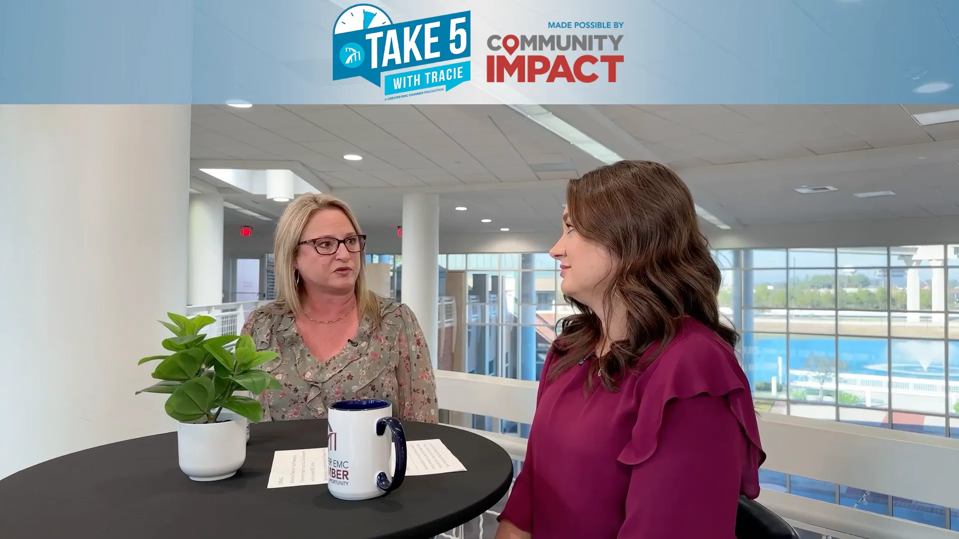 Two women at a small round table in an atrium; one speaking while the other listens, plant and mug visible.