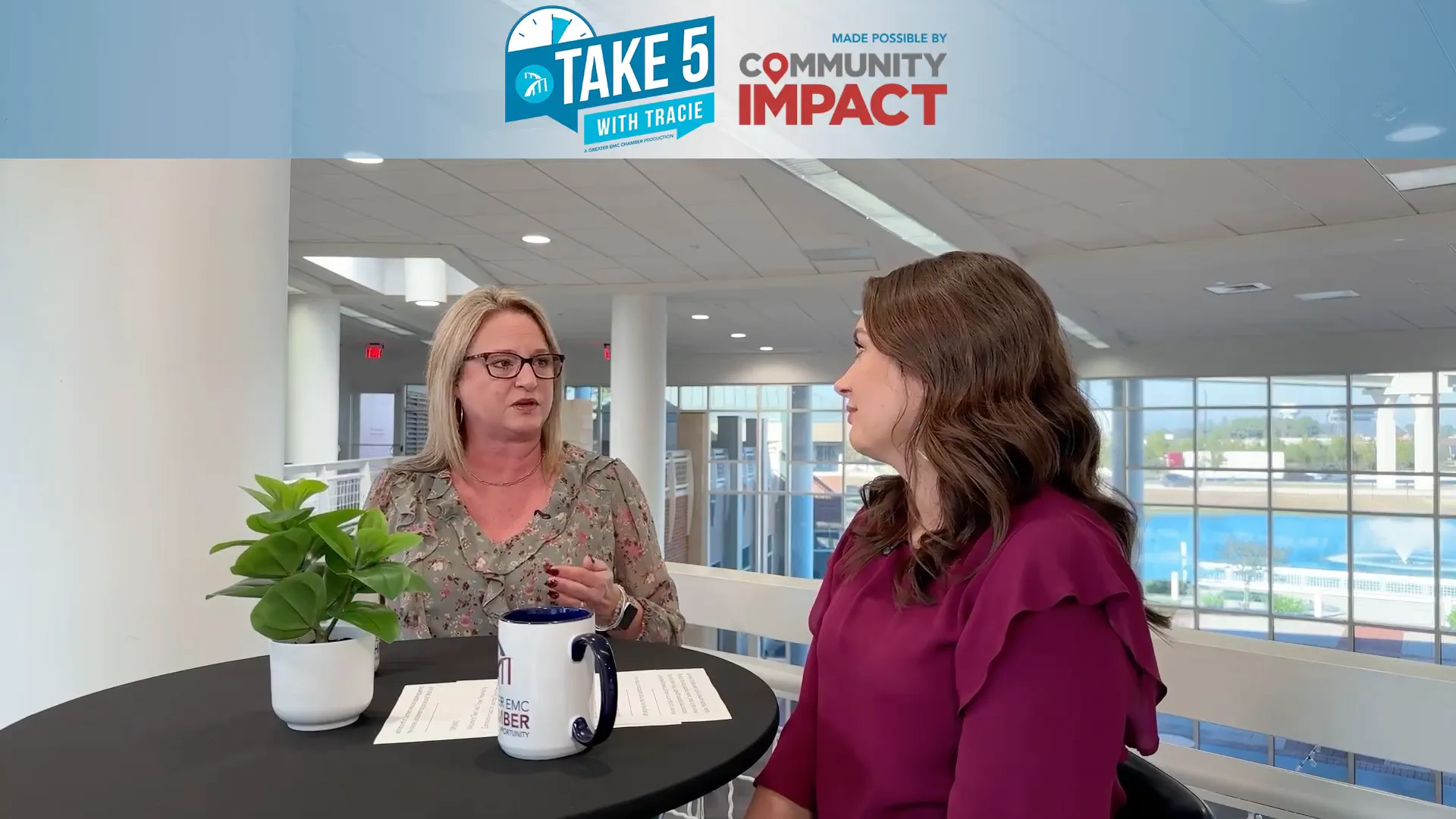 Two people engaged in conversation at a table with a plant and mug, large windows and fountain outside.