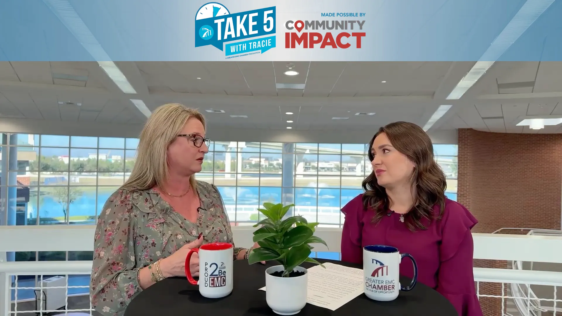 Two women in an interview setting at a round table with coffee mugs and a small potted plant, speaking across from each other in a bright atrium