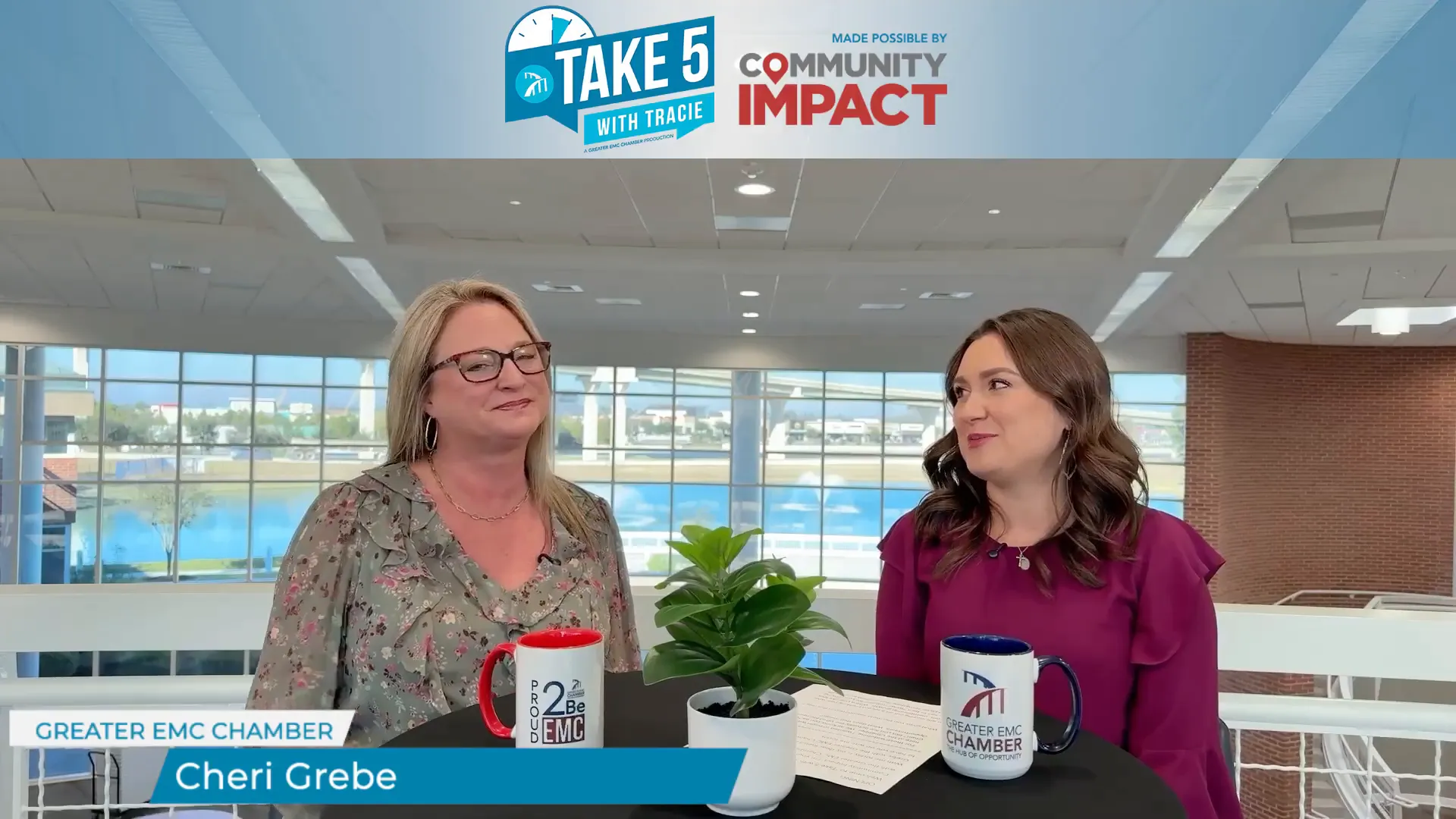 Two women seated at a round table in a bright atrium during an interview, with mugs and a small potted plant on the table and a name lower-third visible.