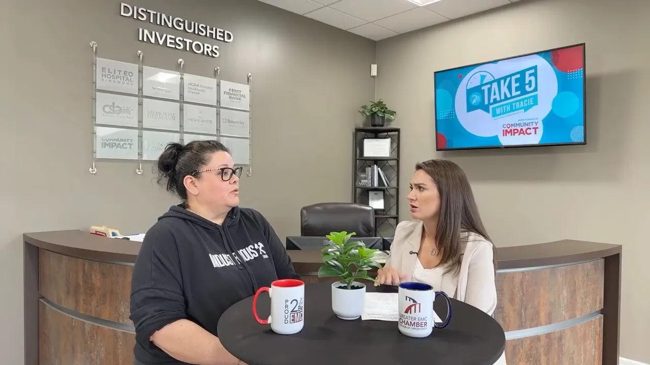 Two women in conversation at a table with a wall-mounted screen displaying Take 5 with Tracie