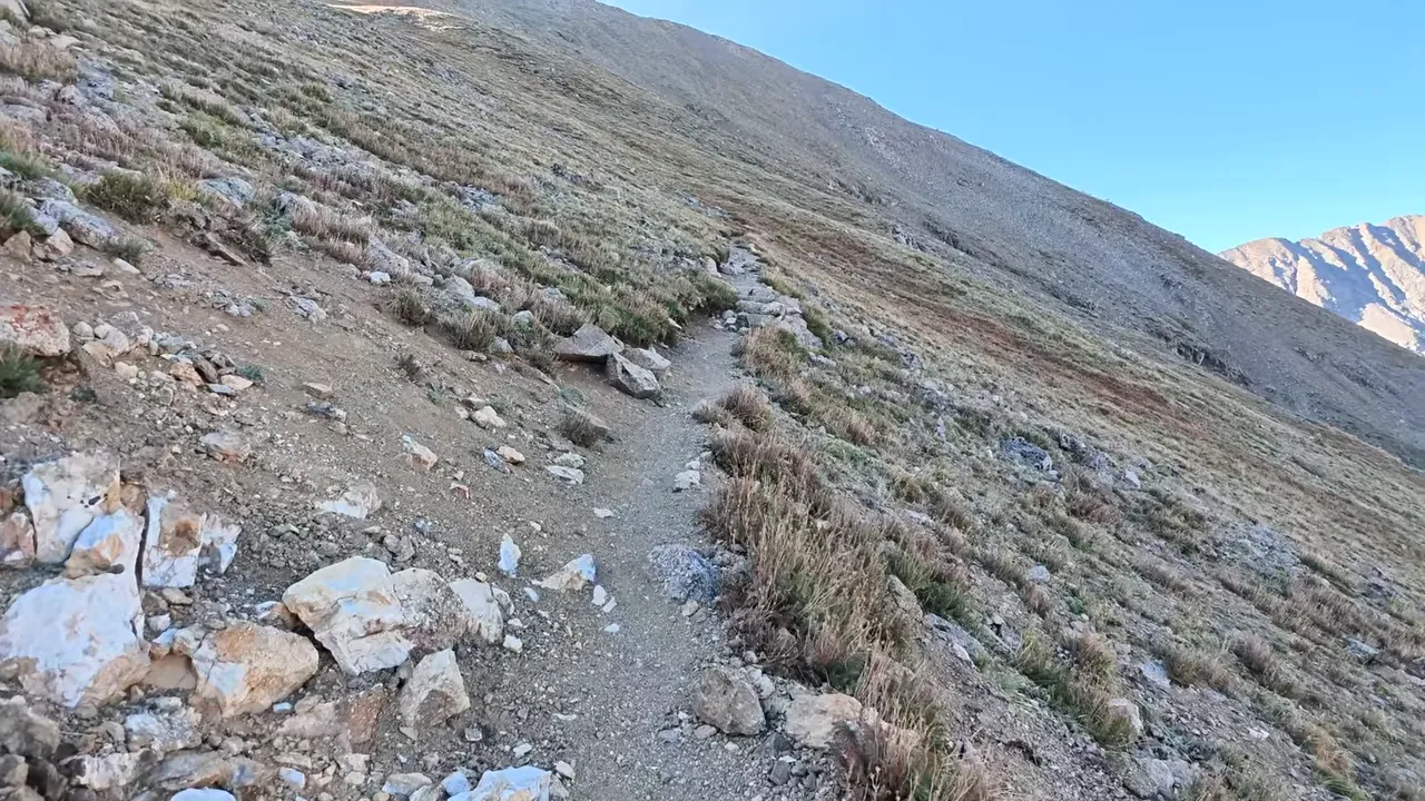 Narrow dirt trail crossing a steep mountainside with loose rocks and sparse grass