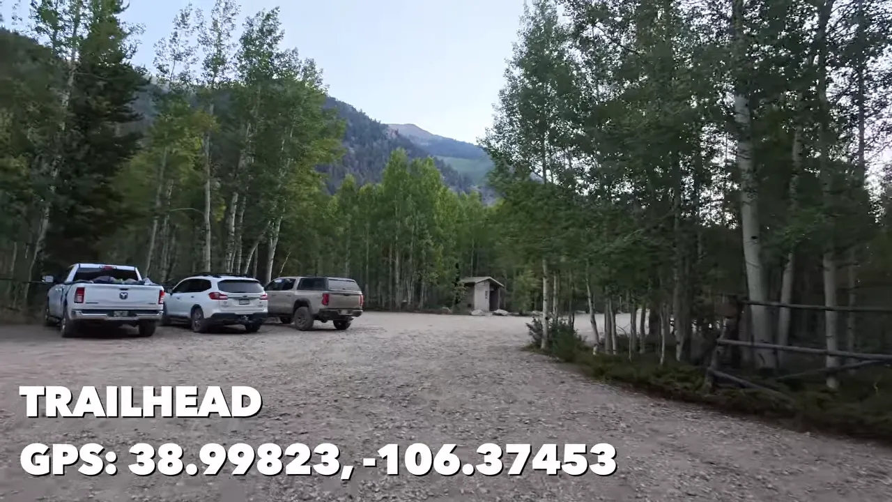 Gravel trailhead parking lot with parked vehicles, a toilet building, and GPS coordinates on screen