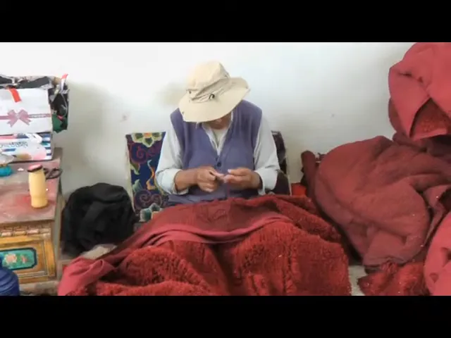 Artigiano tibetano mentre lavora alla realizzazione di un tappeto rosso fatto a mano, circondato da materiali.