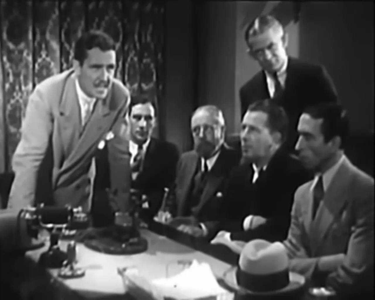 Gang tribunal scene with multiple men at a table, one man leaning forward to speak
