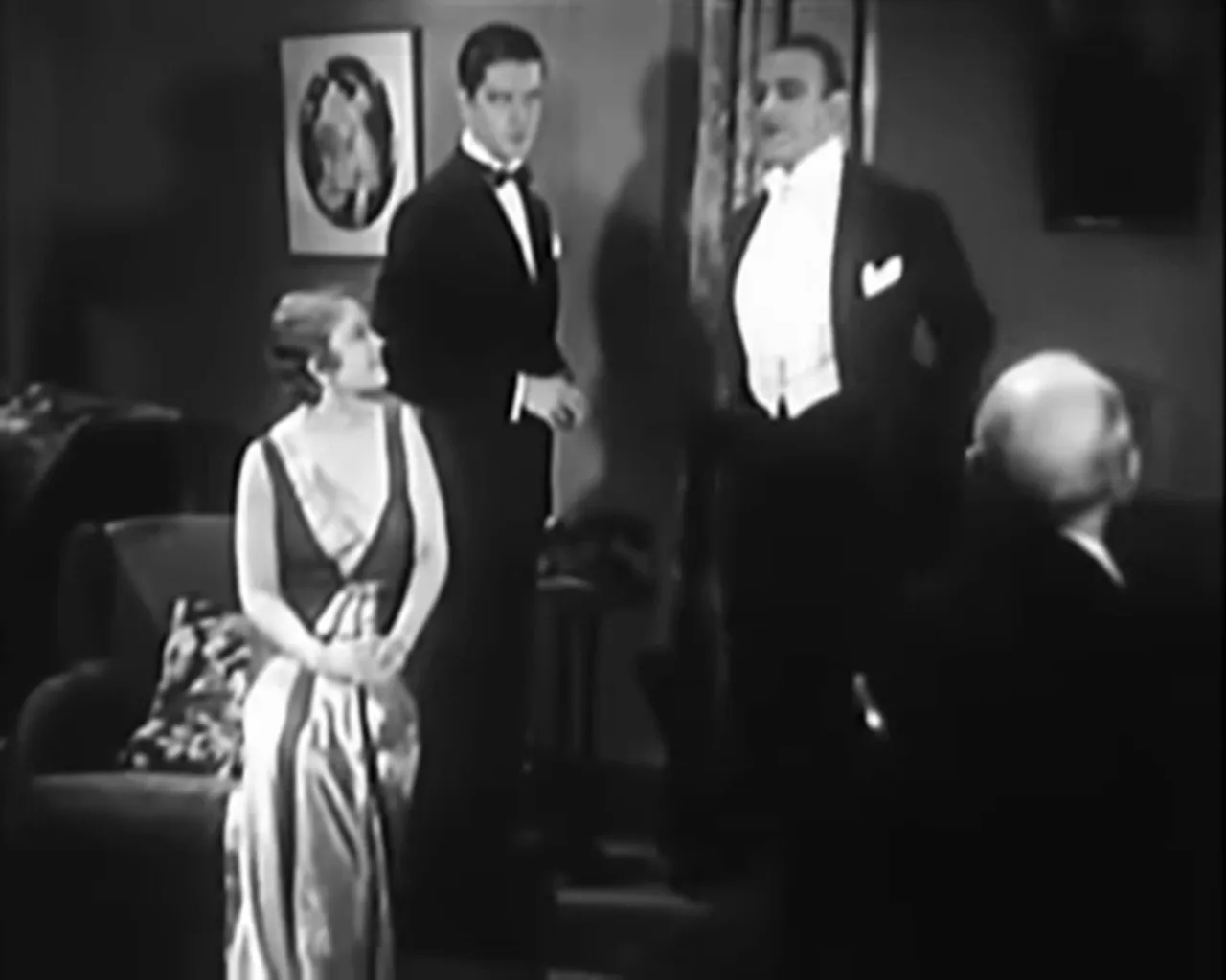 Group scene in an elegant apartment: a seated woman, two standing men in tuxedos, and an older man in the foreground, black-and-white still.
