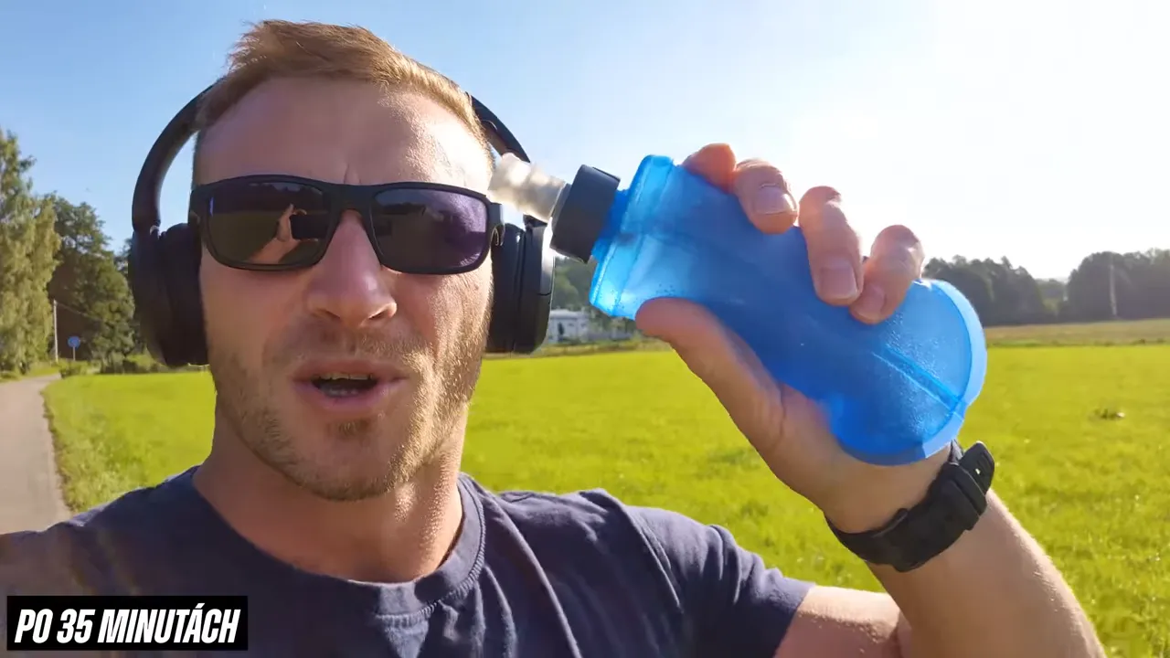Trainer holding a blue handheld water bottle up to camera while running in a sunny field, wearing sunglasses and headphones.