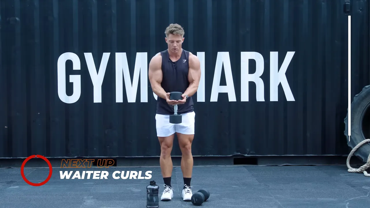 Instructor standing full-frame in front of Gymshark lettering holding a single dumbbell at the head in a close-grip waiter curl position